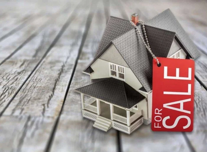 Miniature model of a house with a sale tag hanging from the roof, placed on a wooden surface.