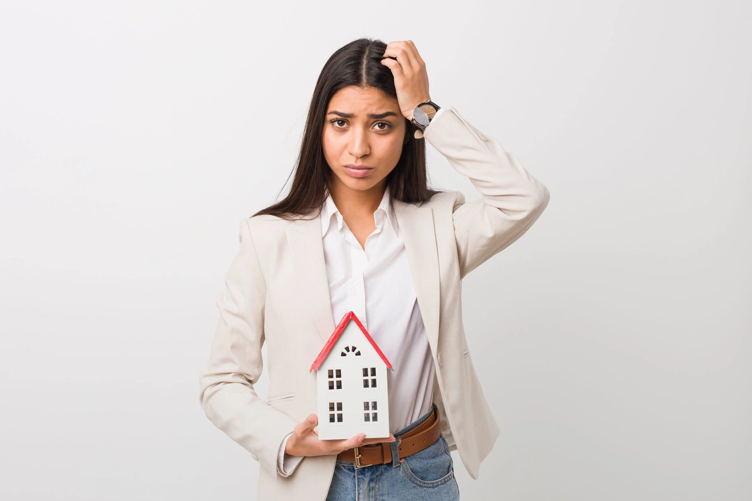 A woman in a beige blazer and white shirt holding a small house model with a red roof, looking confused and touching her head.