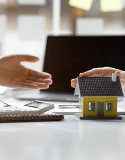 Two people exchanging a handshake in an office with a small yellow model house on the table.