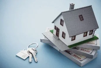 A miniature house model resting on top of a stack of dollar bills, with a key and keychain placed beside it.