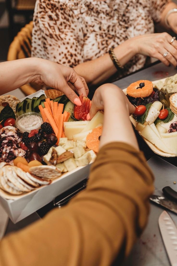 Two people reaching for a large cheese and fruit platter with vegetables, cheese, crackers, grapes, berries, cherry tomatoes, and a small container of dip.