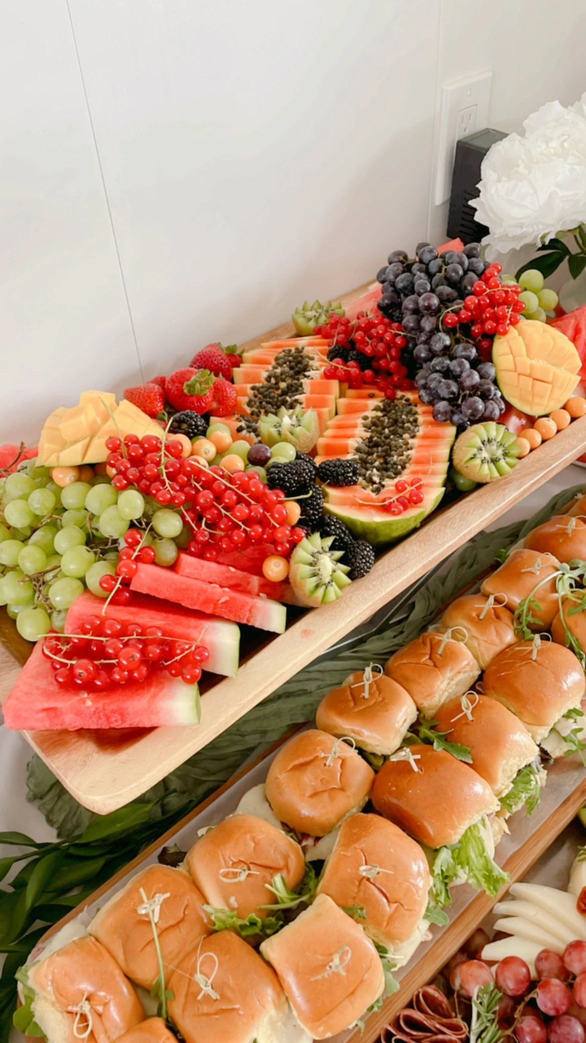 Display of various fresh fruits including watermelon, green and red grapes, strawberries, blackberries, kiwis, cantaloupe, and papaya arranged on wooden platters, along with sliders on buns with lettuce and tied with toothpicks.