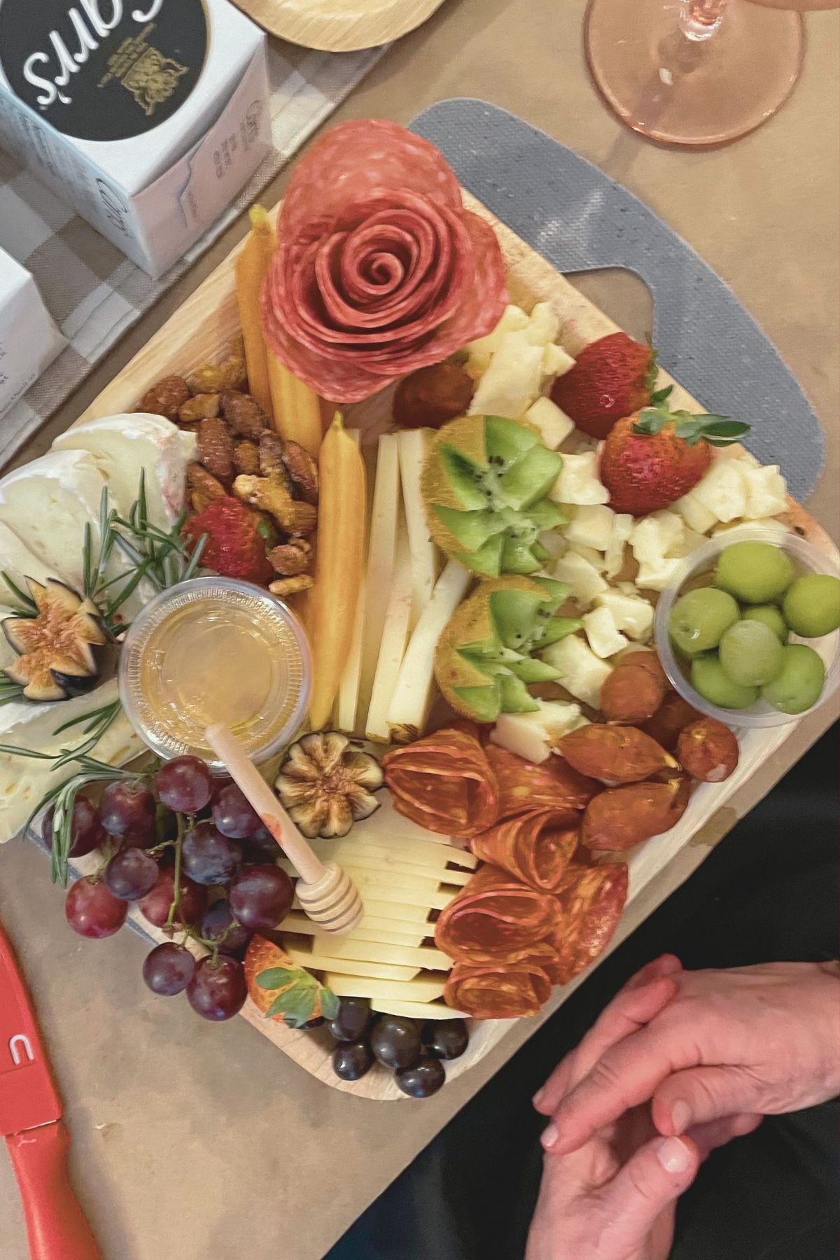 A cheese and charcuterie board with cured meats, various cheeses, fresh fruits like strawberries, kiwifruit, grapes, figs, strawberries, and nuts, alongside condiments and a honey dipper, arranged on a wooden board.