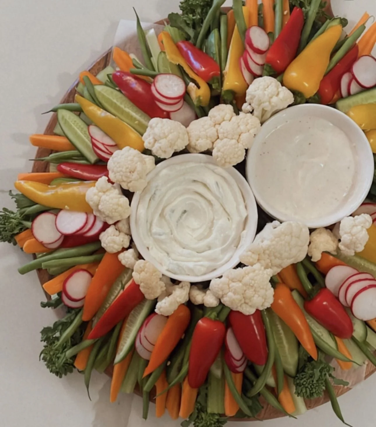 A colorful platter of fresh vegetables including cauliflower, radishes, cherry peppers, cucumber, carrots, and yellow peppers, with two bowls of creamy dips in the center.