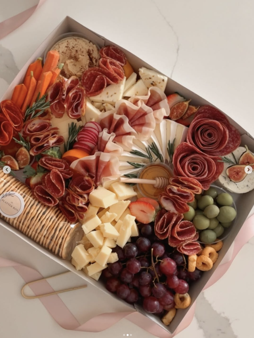 Cheese, grapes, cured meats, crackers, strawberries, figs, carrots, radishes, and herbs arranged in a serving box.