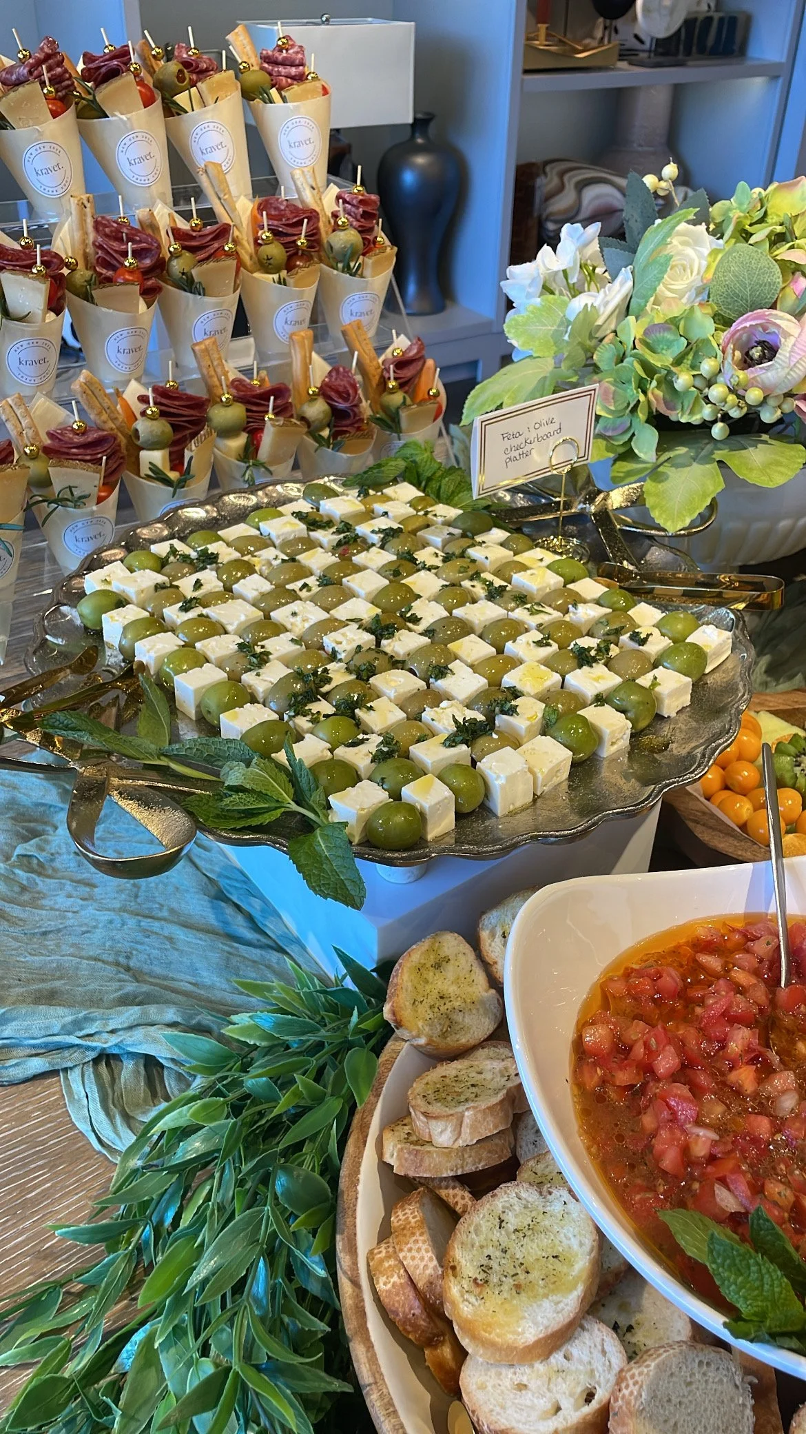 A table with an assortment of appetizers including cheese and grapes, a bowl of tomato-based dip with bread slices, and skewers with cured meat, cheese, and olives. There are also fresh green leaves and a flower arrangement for decoration.