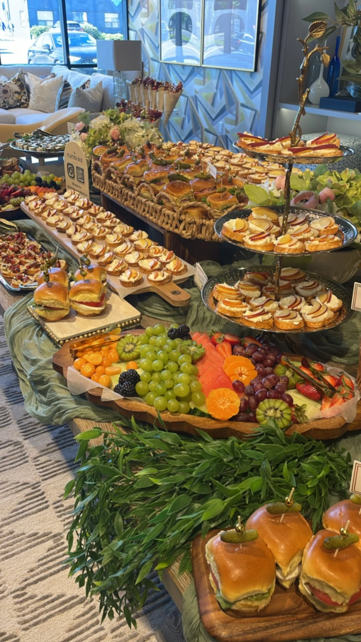A buffet table with assorted finger foods, miniature sandwiches, fresh fruit, and desserts at a social gathering or event in a bright, modern room.