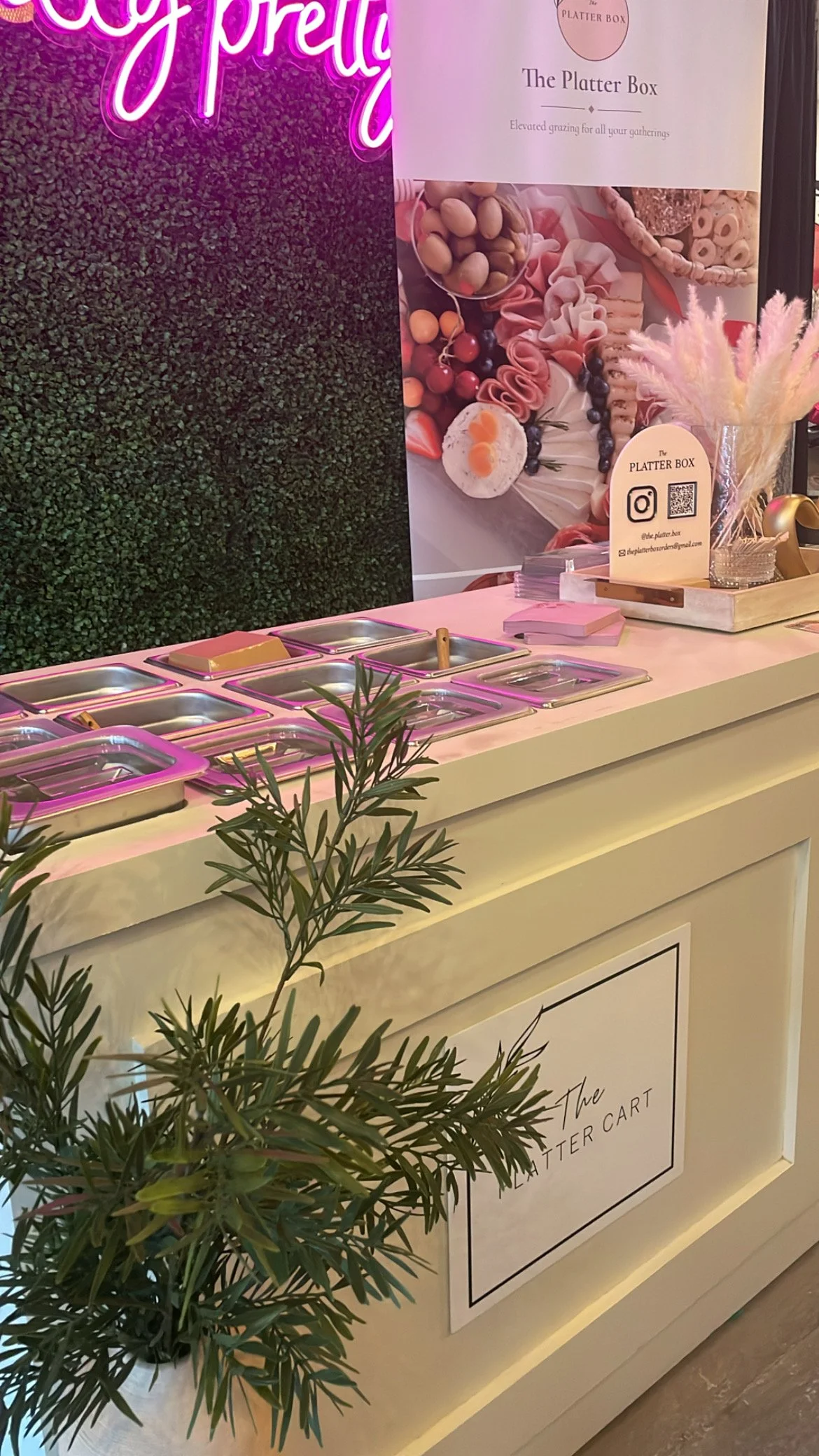 A white counter with empty serving trays, pink napkins, and a small plant in the foreground. There is a sign for 'The Platter Box' with a pink floral background behind it, decorated with a pink and white feather arrangement.