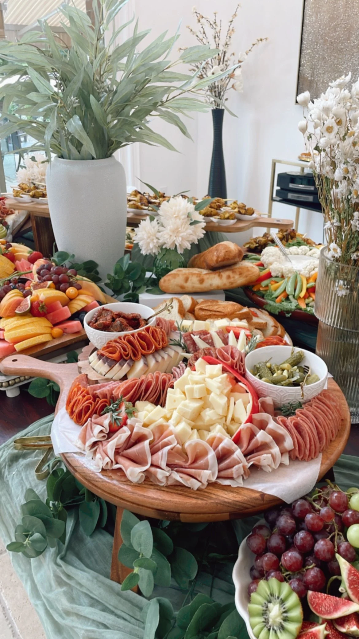 A buffet table with a variety of fruits, cheeses, meats, and bread, decorated with large green leaves and white flowers. There are plates of sliced fruits, grapes, and a cheese platter, with a pitcher of flowers and bread loaves in the background.