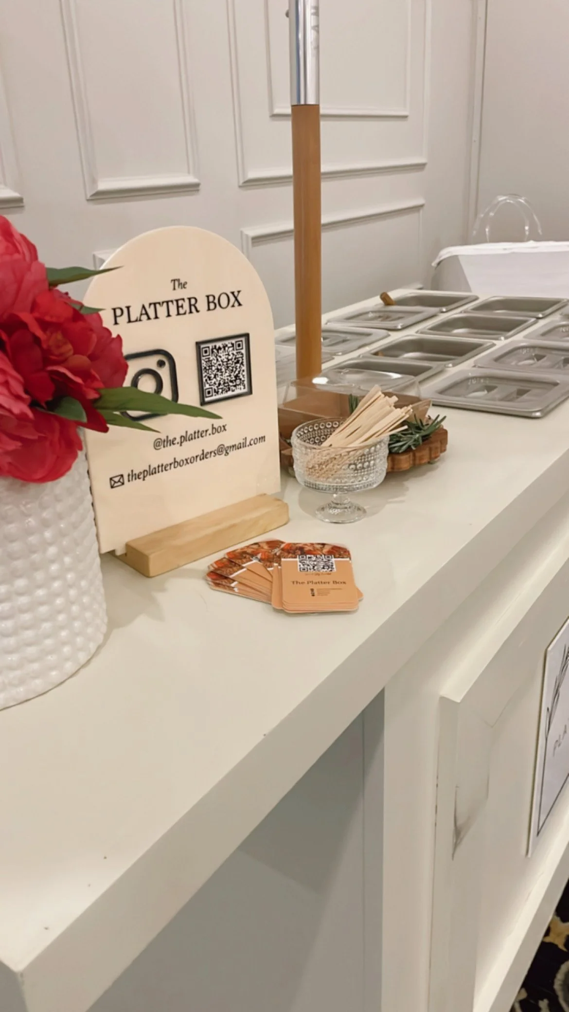 A white table displaying a sign for 'The Platter Box' with contact information, a QR code, and social media handle. There are also several empty food containers, a small bowl with paper napkins, a flower arrangement in a white textured vase, and a rack with silver lids for food.