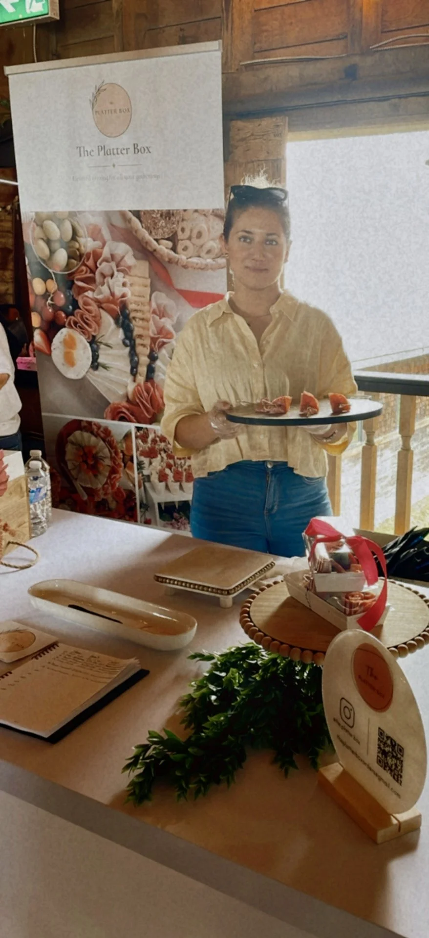 A woman holding a black tray with small appetizers in a rustic venue with wooden walls and decor featuring a promotional banner for The Platter Box.