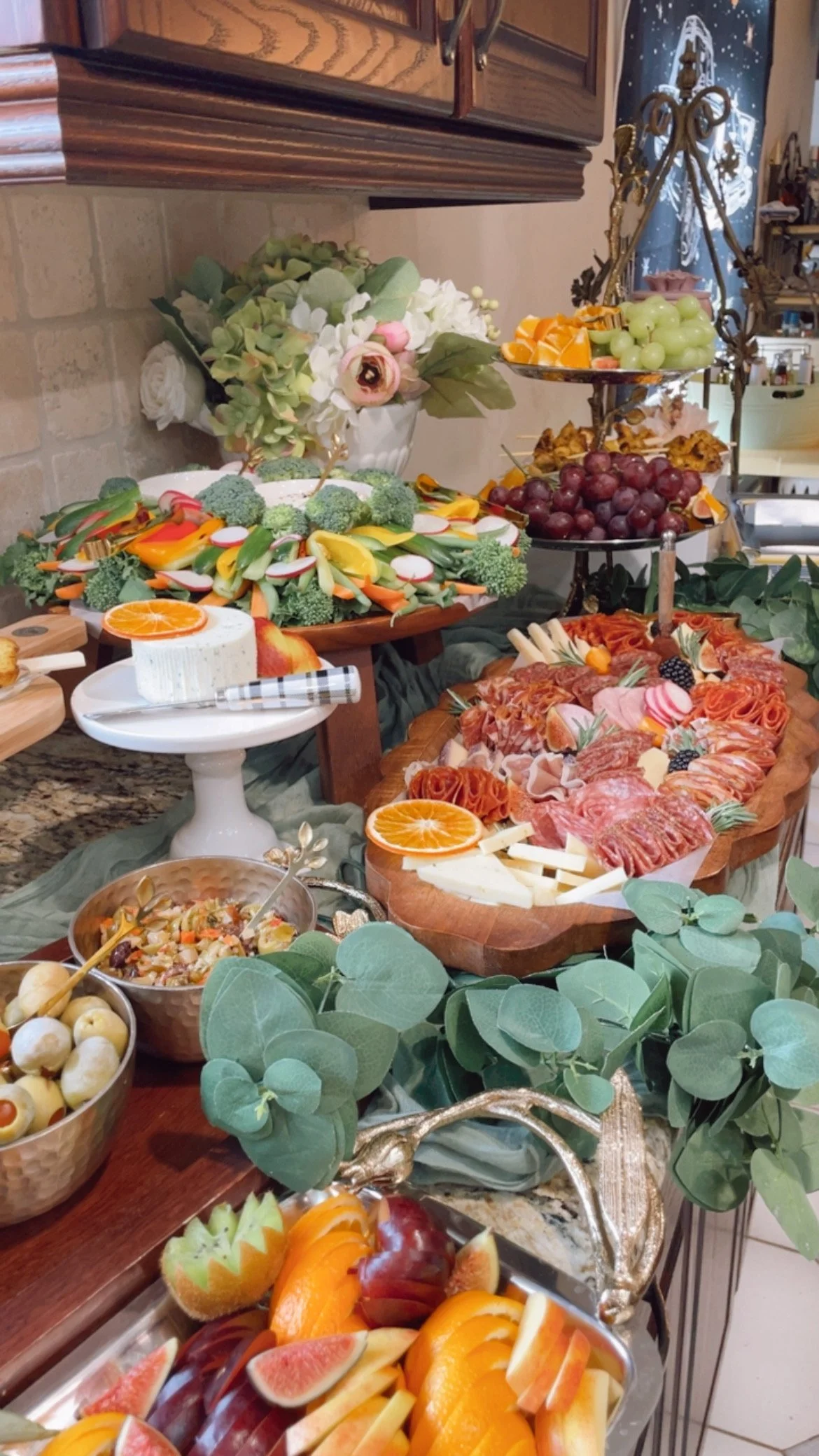 A festive holiday food display with an assortment of fruits, vegetables, cheese, cold cuts, and a flower arrangement on a table decorated with greenery.