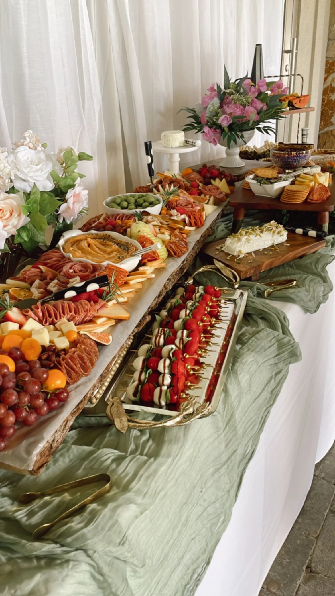A buffet table with various appetizers and finger foods, including cheese, meats, fruit, and desserts, decorated with floral arrangements and greenery.