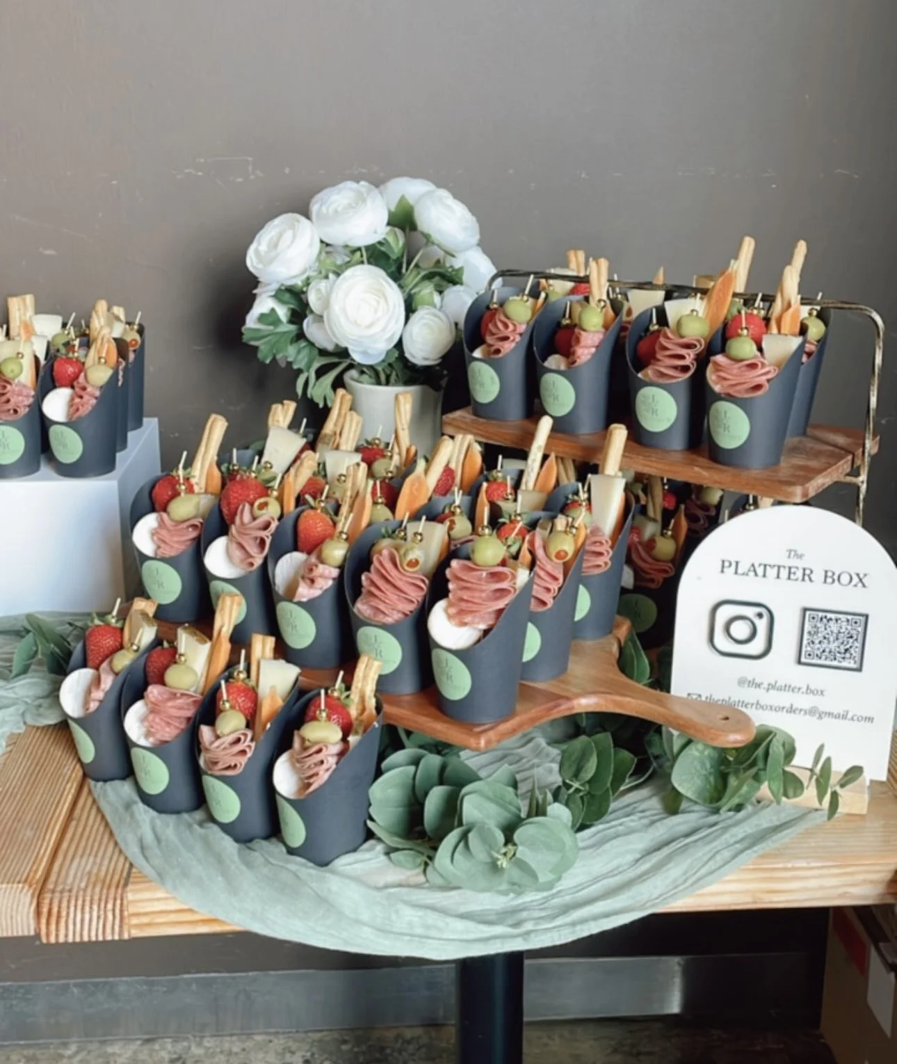 Display of small snack cones filled with cured meat, cheese, strawberries, green grapes, and breadsticks, arranged on a wooden tiered stand with a floral arrangement of white roses and greenery nearby.