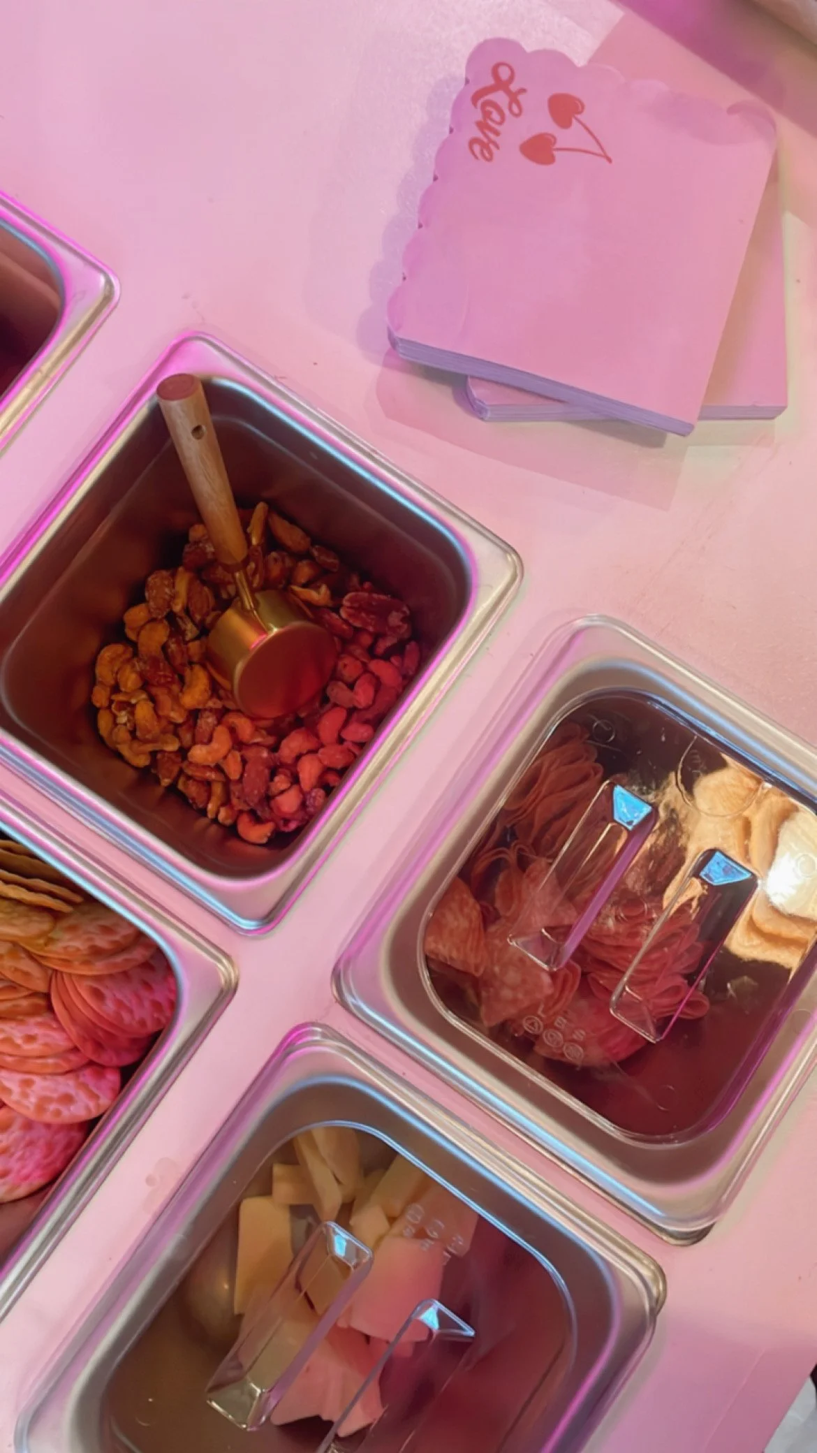 Pink tables with stainless steel containers filled with different types of food, including nuts, cold cuts, and cheeses. Pink napkins with a heart and arrow design are stacked on the table.