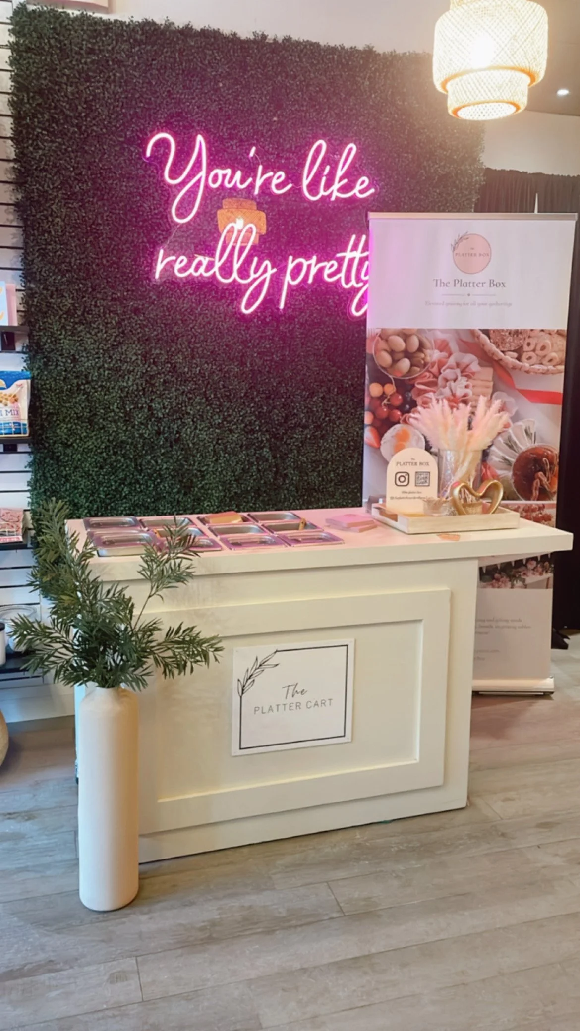 A white counter with a potted plant in a white vase, a sign that says "The Platter Cart," and various pink-colored dishes. Behind the counter, there is a green textured wall with a neon sign that reads "You're like really pretty" in pink. To the right, there is a banner with the logo and name "The Platter Box," showing images of assorted snacks and a QR code.