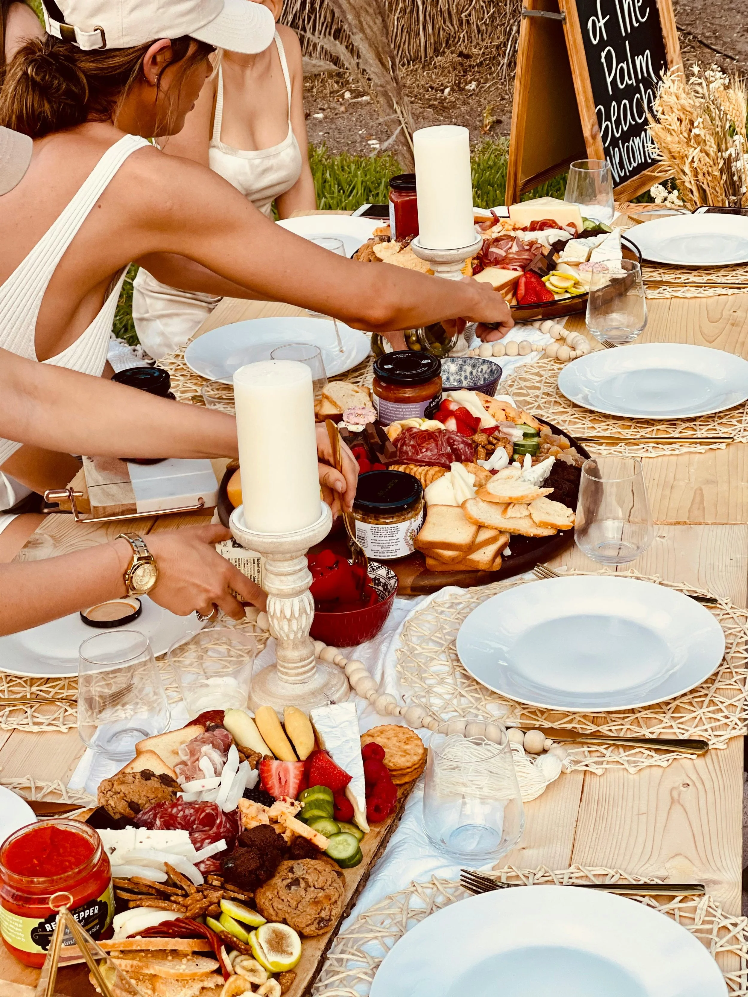 A table set for an outdoor meal with a cheese, meat, vegetable, and fruit platter, plates, glasses, and candles. Two women are reaching for food on the platter. A chalkboard sign reads 'of the Palm Beach welcome'.