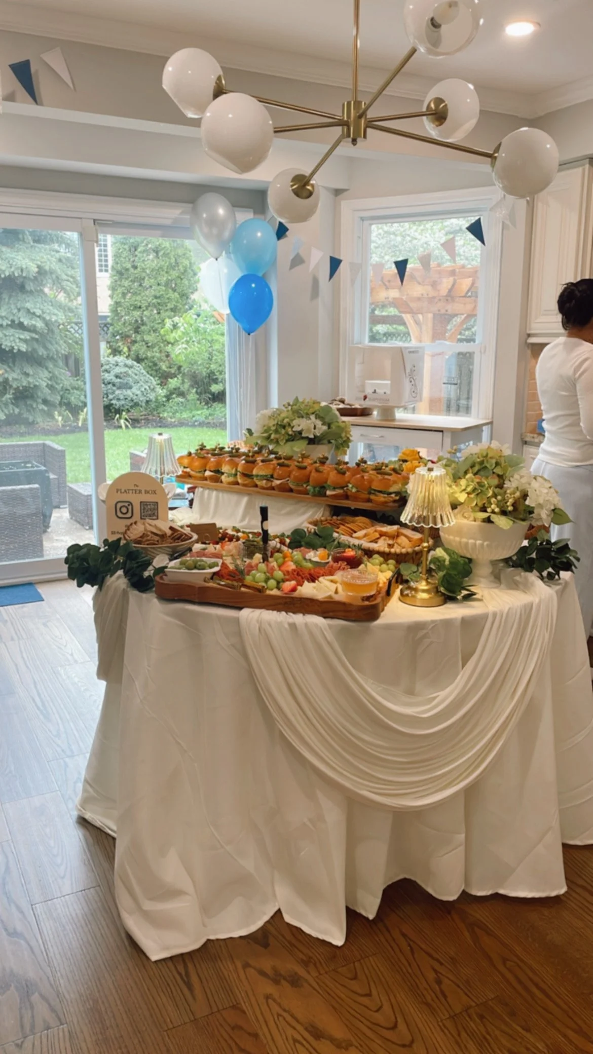 A festive indoor setup with a decorated table of food, balloons, and white draped tablecloth, near large windows and a garden view.