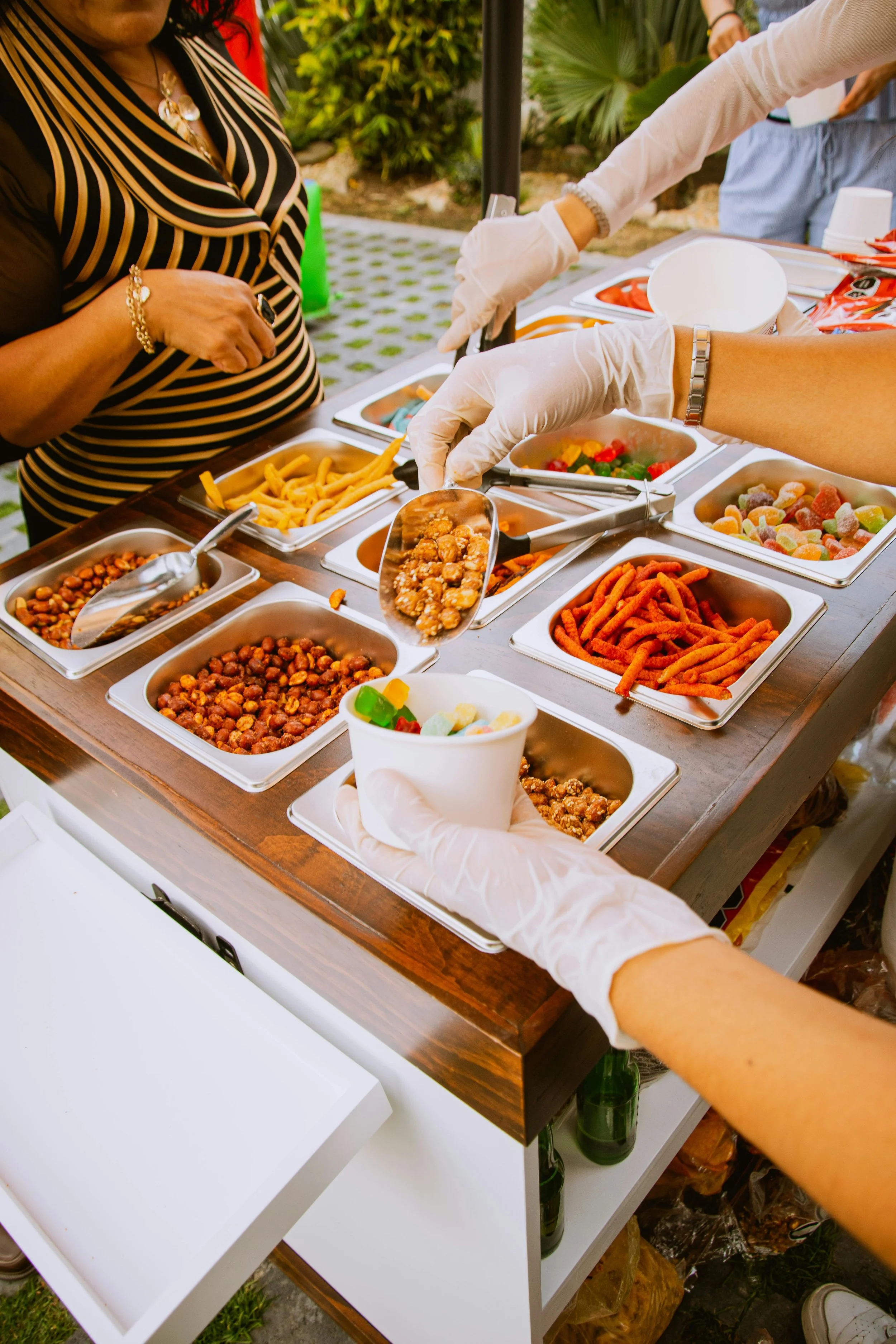 A buffet table with various bowls of snacks and candies, with people serving themselves, outdoors in a garden or park setting.