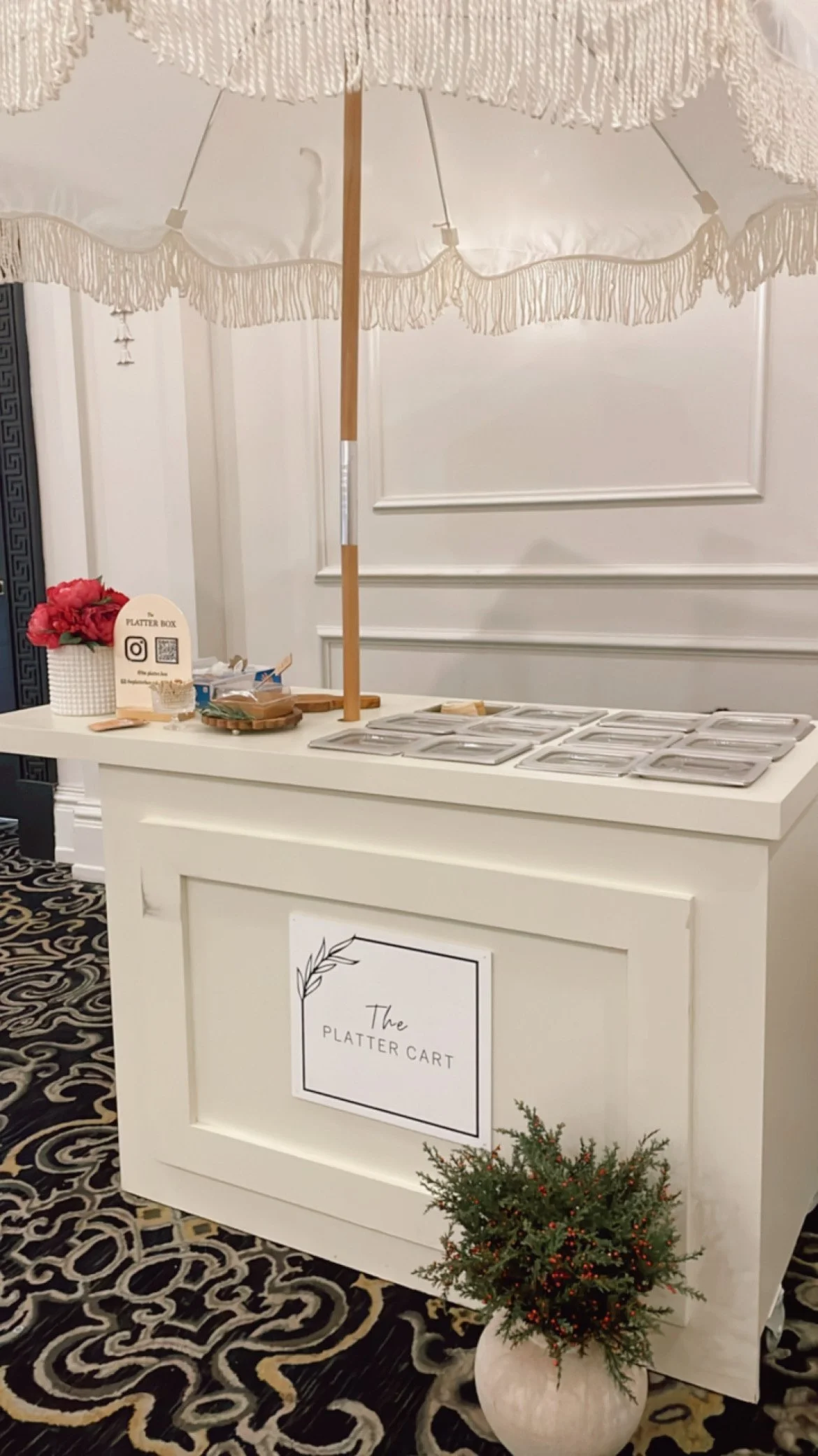 A white counter with a sign reading 'The Platter Cart' and small trays, a small potted plant with red berries in front, a white vase with red flowers, and a large white fringed umbrella above.