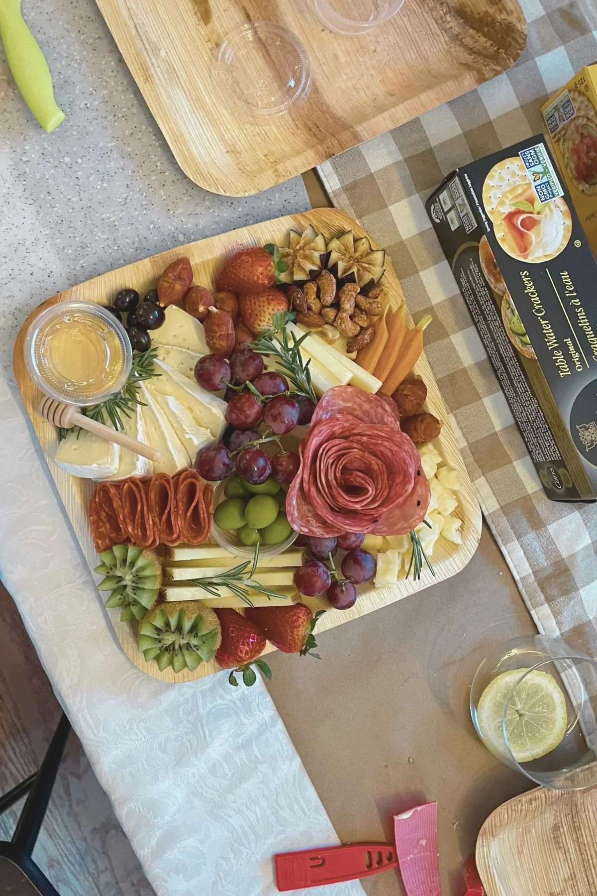Charcuterie board with assorted cheeses, grapes, strawberries, kiwi, ham, salami, dried fruits, and herbs, placed on a table with lemon drinks nearby.
