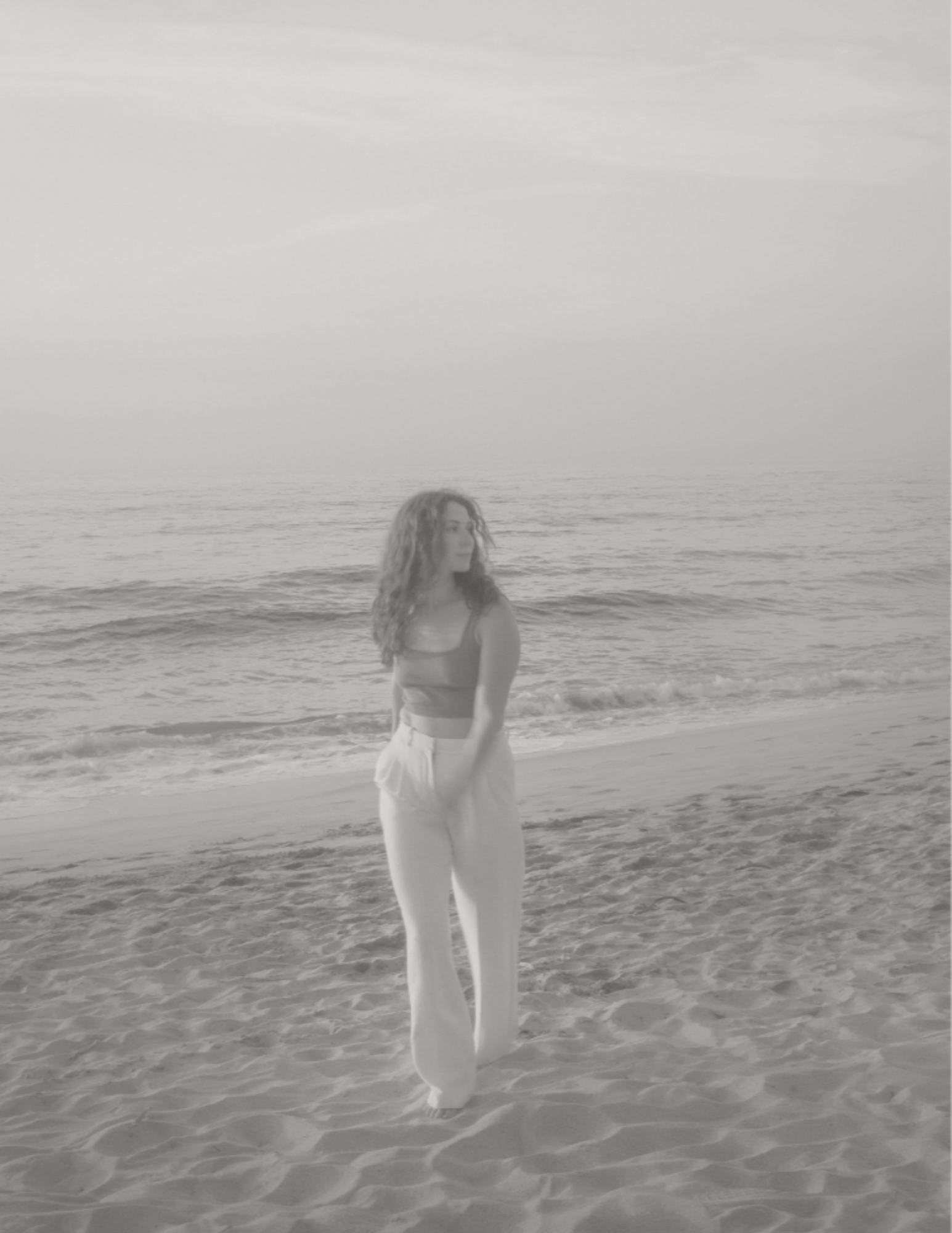 A woman with curly hair walking on the beach with ocean waves and a cloudy sky in the background.