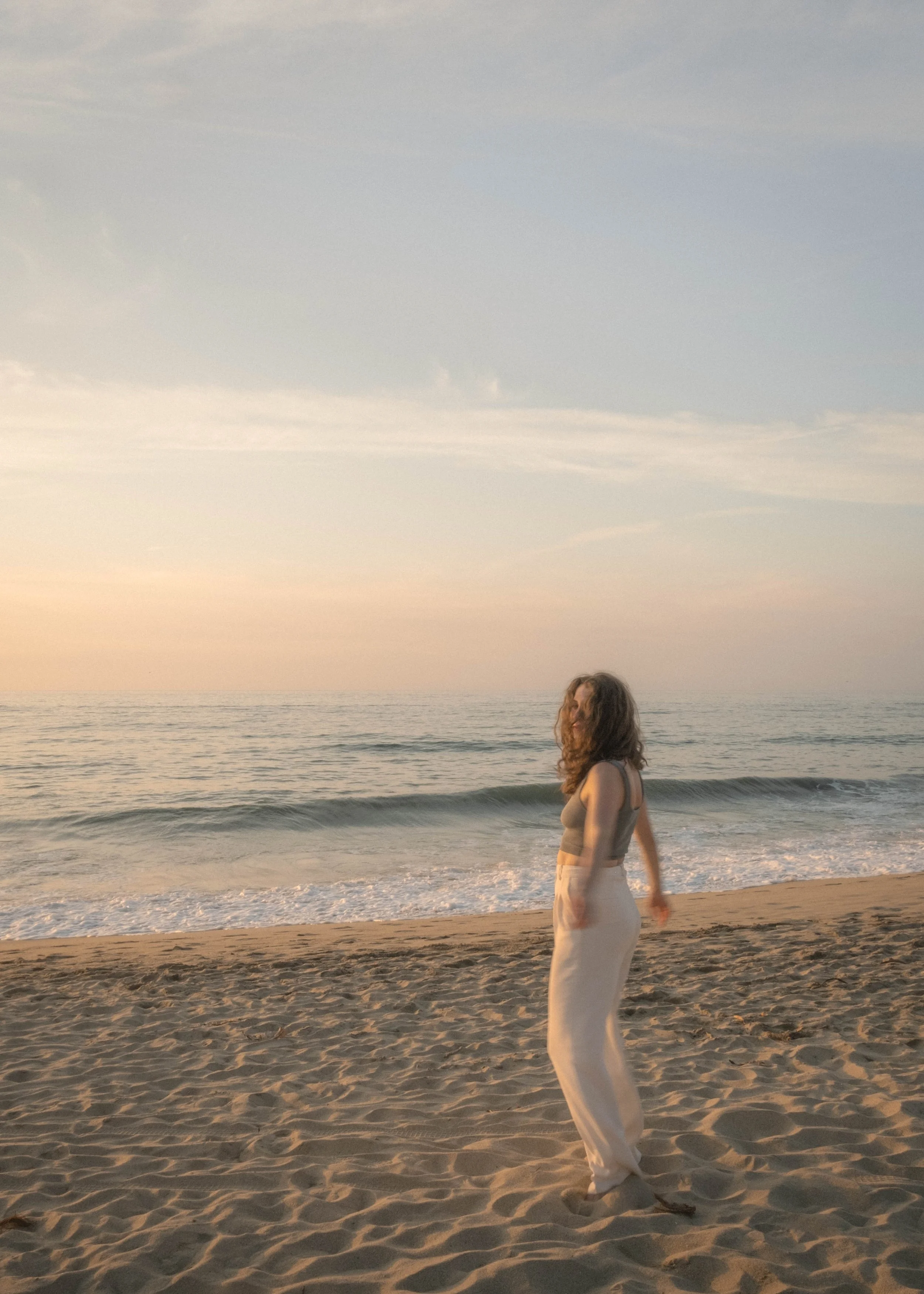 girl with curly hair walking on a serene beach at sunset in Mexico barefoot wearing loose fitted white pants