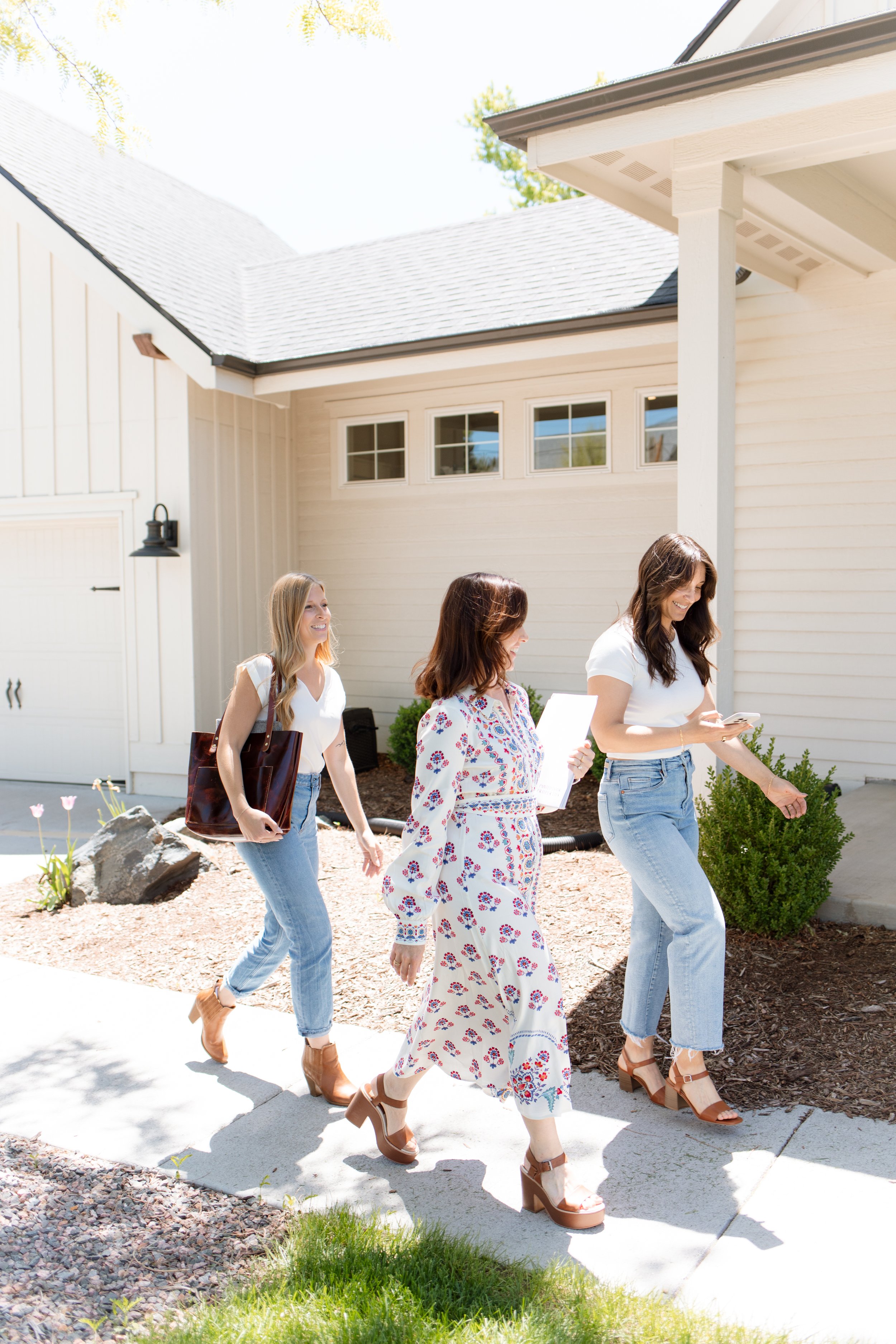Three women walking outside a white house, one holding a tote bag, one holding papers, and one looking at her phone, all smiling.