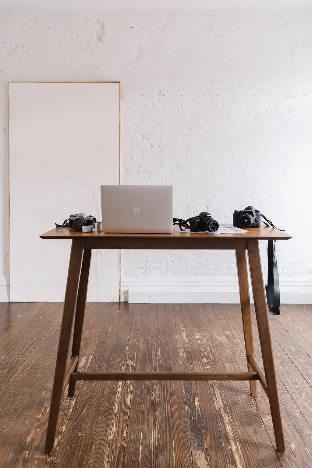 Wooden table with a silver laptop, three cameras, and a lens cap, set against a white brick wall in a minimalist room with wooden floor.