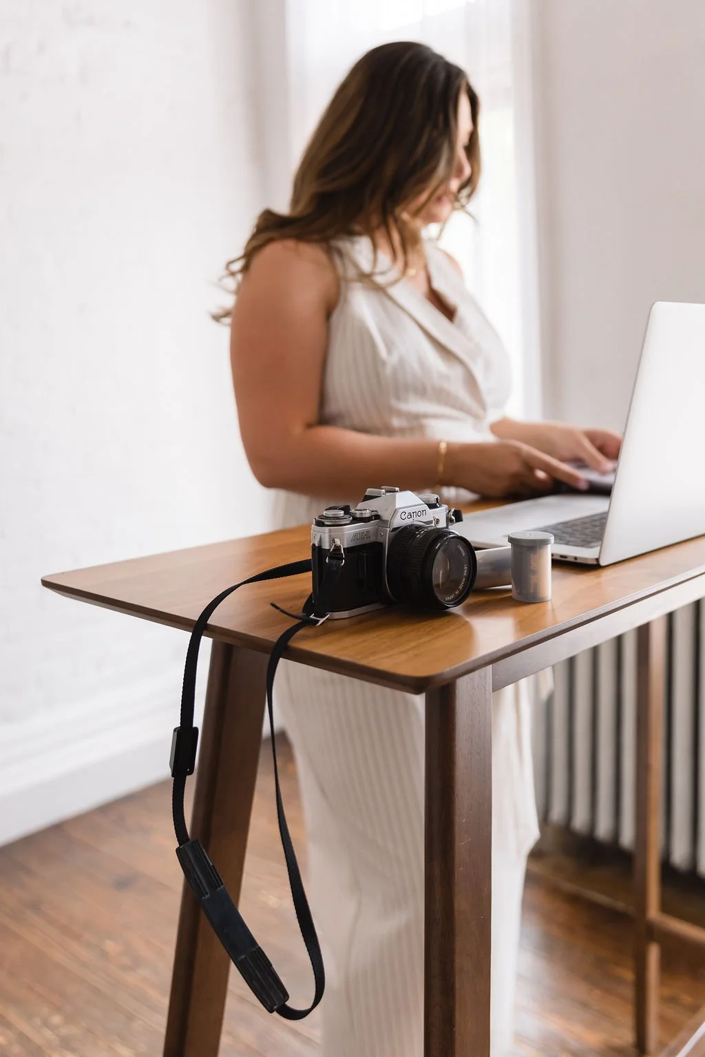 A woman in a white dress working on a laptop at a wooden table. There is a vintage Canon camera and a film roll on the table.