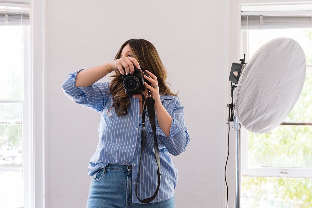 Woman taking a photo with a camera in a room with natural light, standing near a studio light and a window.