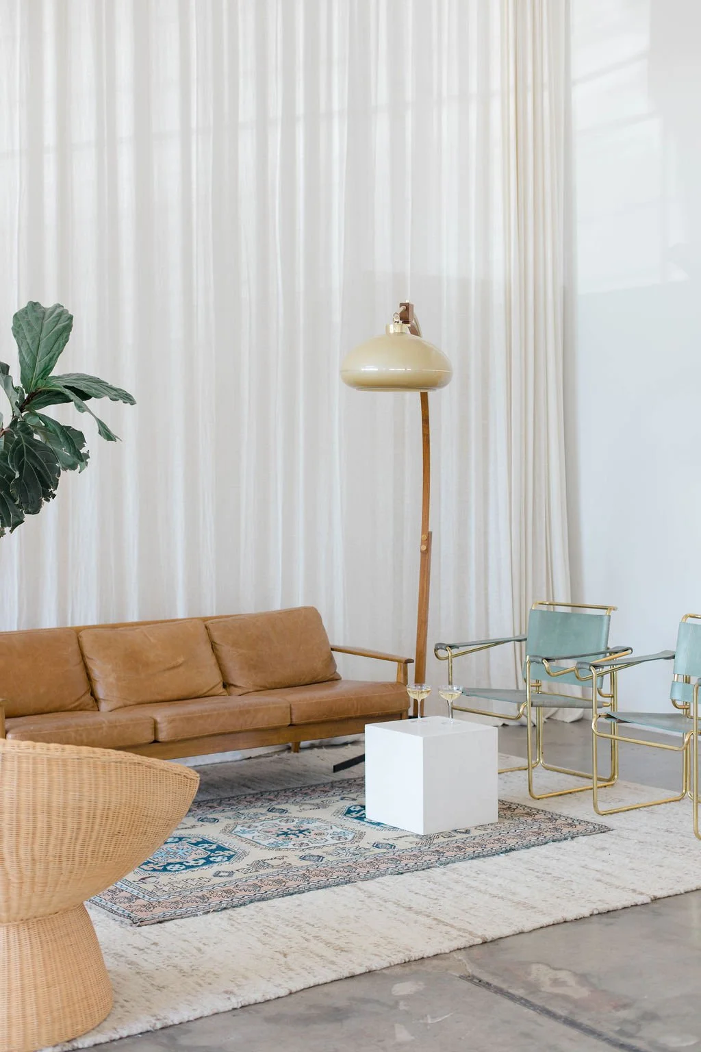 Living room with a brown leather sofa, light green armchairs with gold frames, a large potted plant, a tall floor lamp, a white square side table, a patterned area rug, and floor-to-ceiling white curtains.