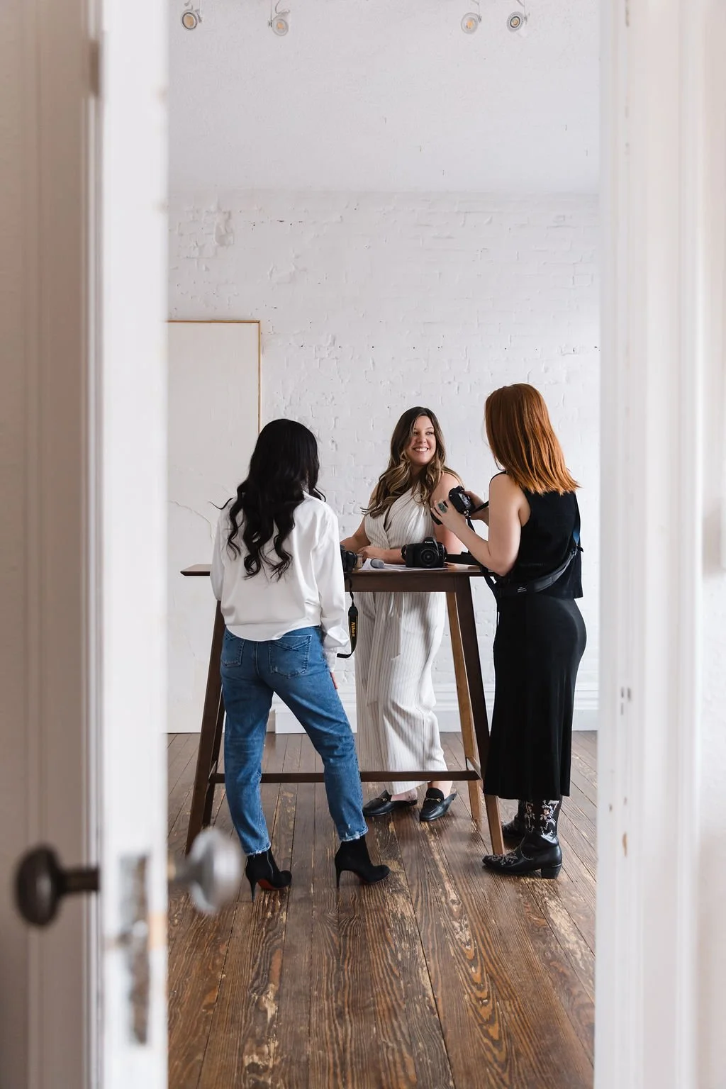Three women in a photography studio, one sitting at a table with cameras, one standing and holding a camera, and one with her back to the camera, all smiling and engaged in conversation.