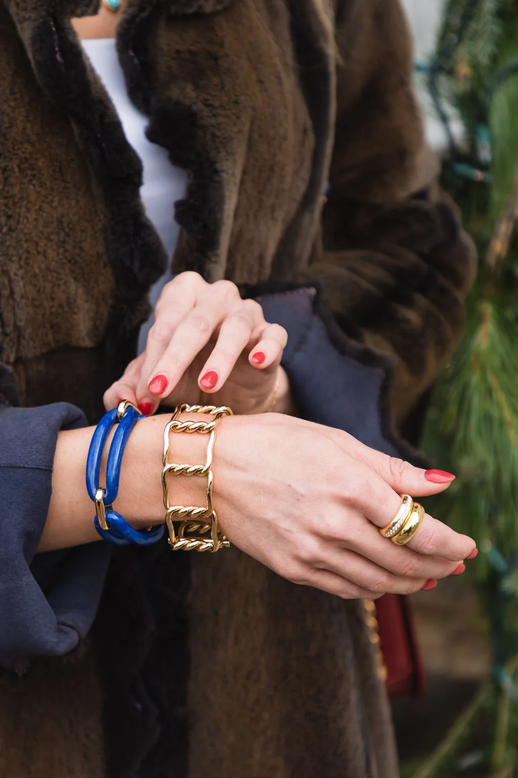 Close-up of two women clasping hands, displaying jewelry including a blue bracelet, a gold chain bracelet, and a gold ring with embedded stones, with one woman wearing a brown fur coat and the other a dark-colored sleeve, against a background of greenery.