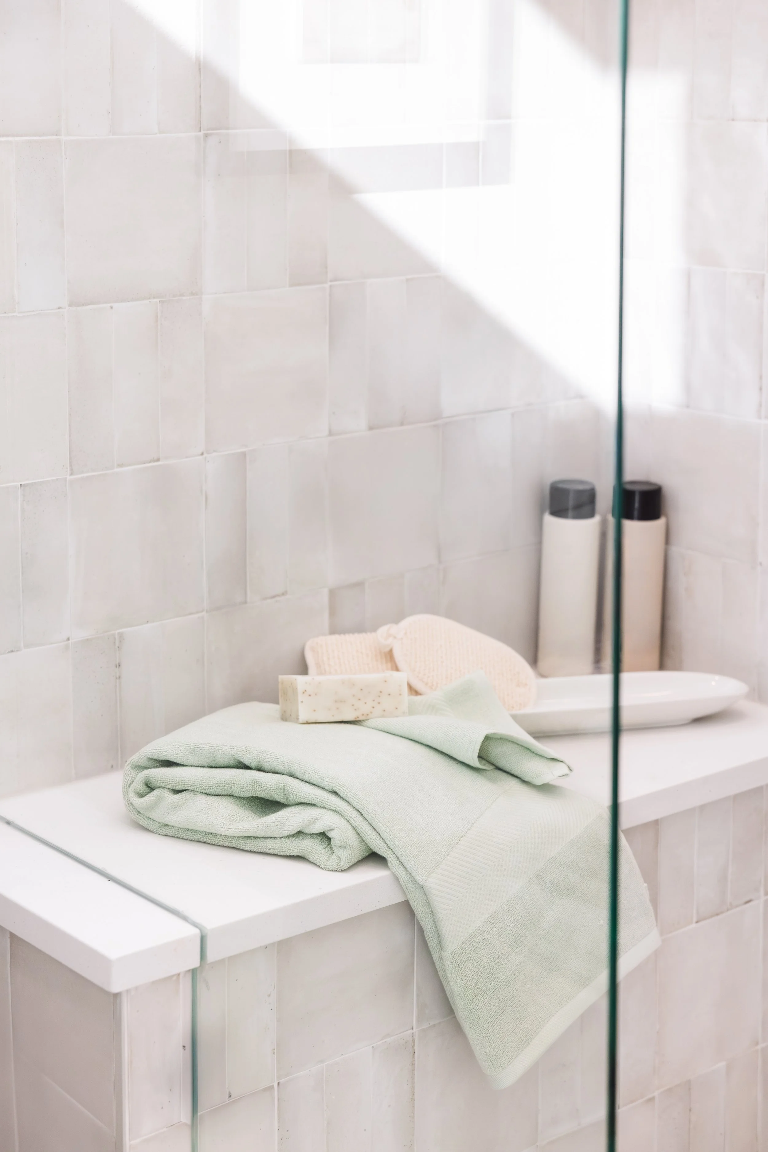 A neatly folded green towel on a bathroom counter with soap, loofahs, and shampoo bottles.