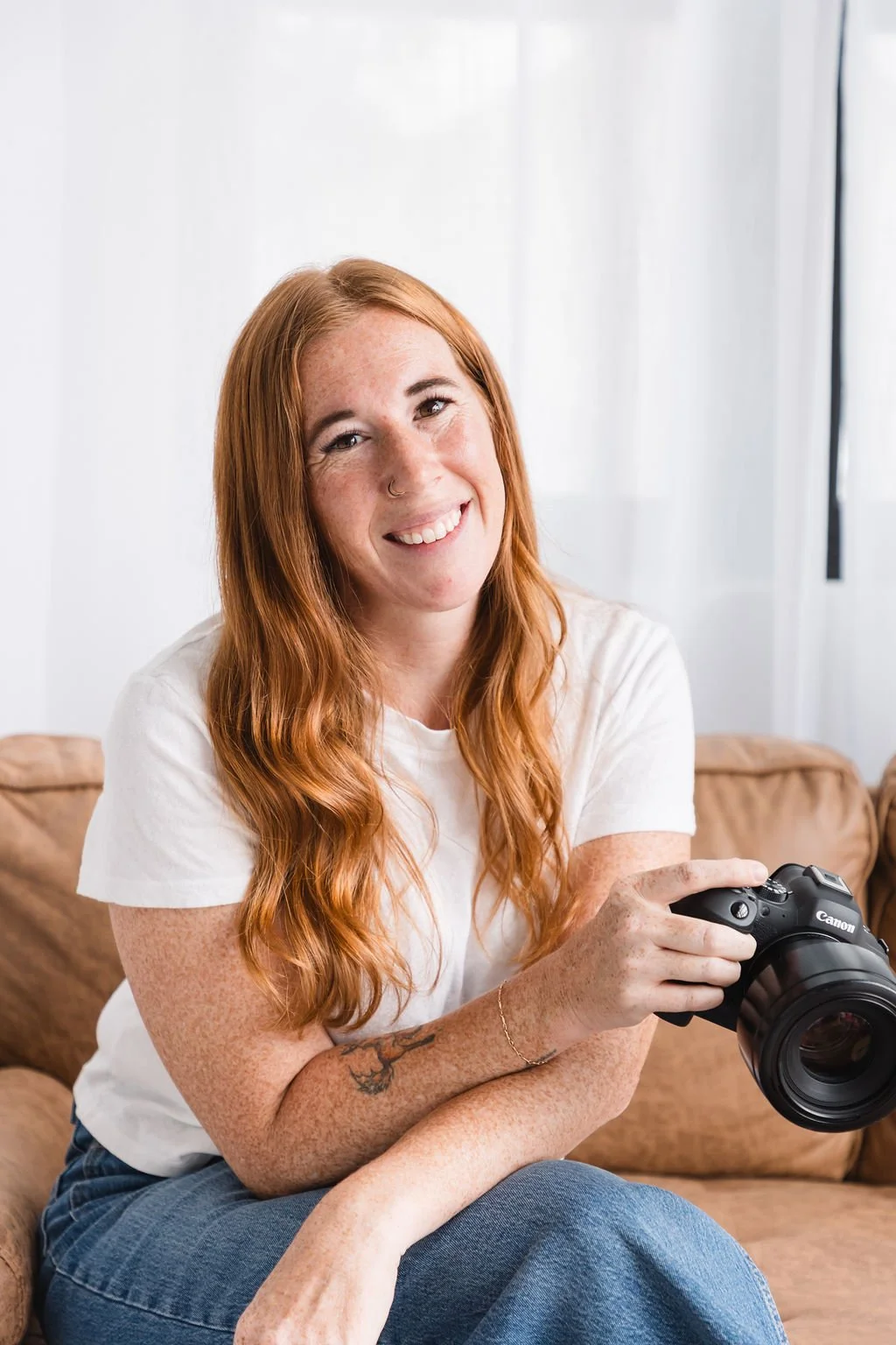 A woman with long red hair, freckles, and a nose ring sitting on a brown couch holding a professional camera, smiling at the camera.