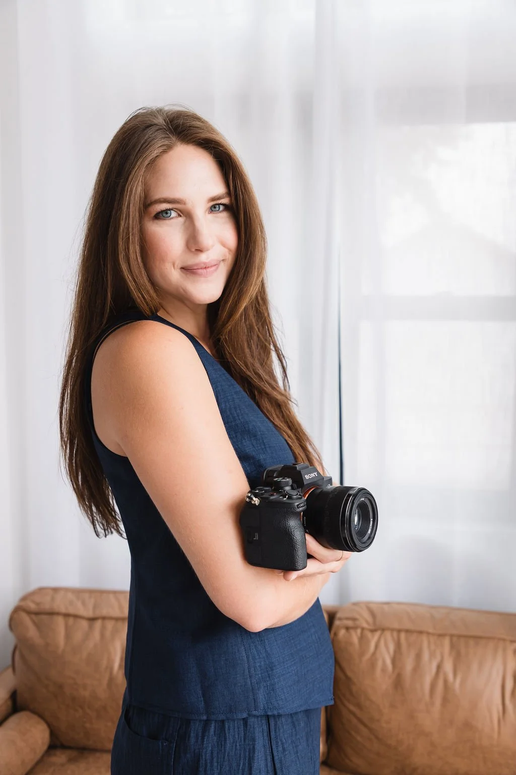 A woman with long brown hair and blue eyes holding a professional camera, standing in a room with a brown sofa and white curtains.