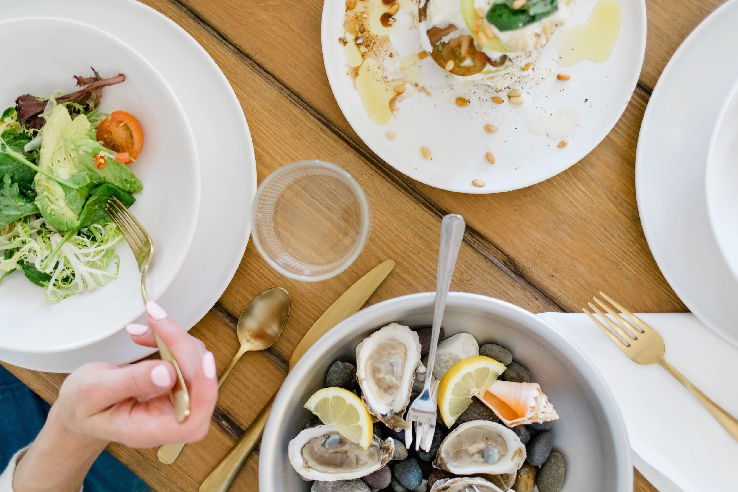 Top-down view of a dining table with various dishes, including a bowl of fresh salad with cherry tomatoes on a white plate, a plate with a dessert topped with whipped cream, nuts, and lemon slices, another plate with oysters on rocks and lemon wedges, and cutlery including gold-colored forks and spoons, a clear glass of water, and a hand holding a gold spoon.
