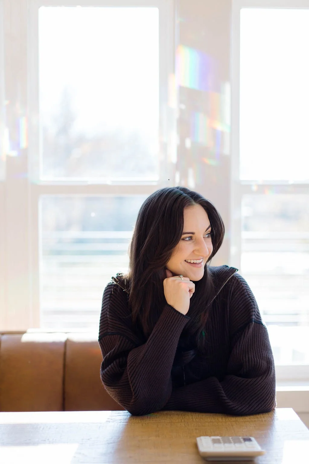 A woman with dark hair smiling, sitting at a wooden table with a calculator in front of her, near large windows with sunlight and rainbow reflections.