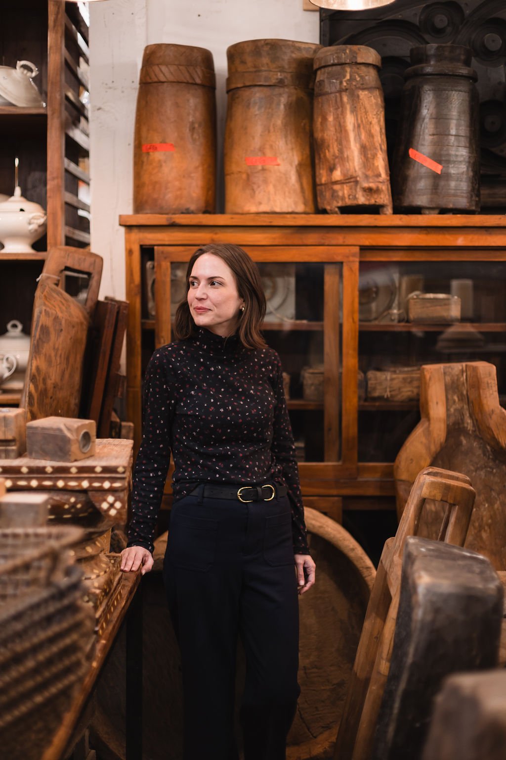 A woman with shoulder-length brown hair wearing a black dress with small pink and white patterns stands inside an antique store, surrounded by wooden furniture and artifacts.