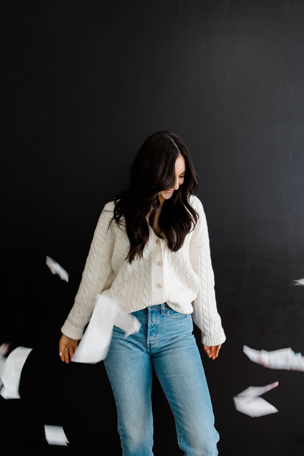 Woman with long dark hair wearing a cream-colored sweater and jeans standing against a black wall with paper flying around.