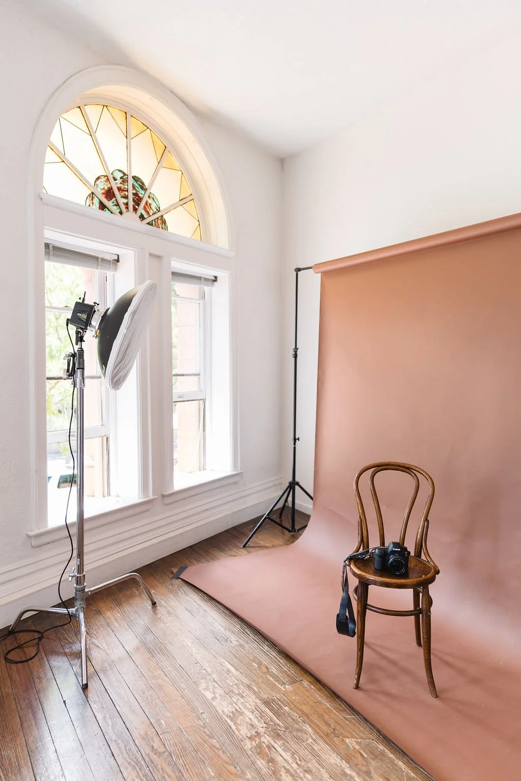 Photography setup with a brown paper backdrop, camera on a chair, studio light, and large windows with stained glass.