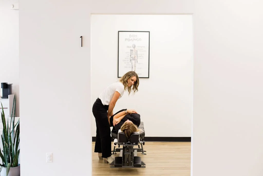 A chiropractor adjusting a patient's neck in an examination room, with a medical chart on the wall.