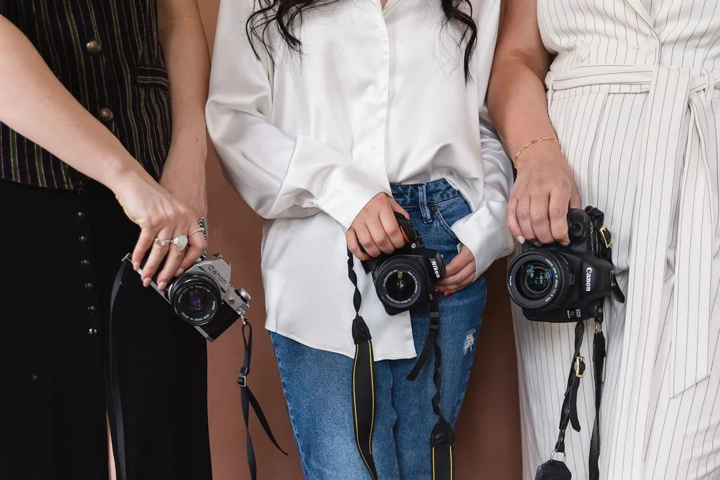 Three women holding cameras, standing side by side against a neutral background.