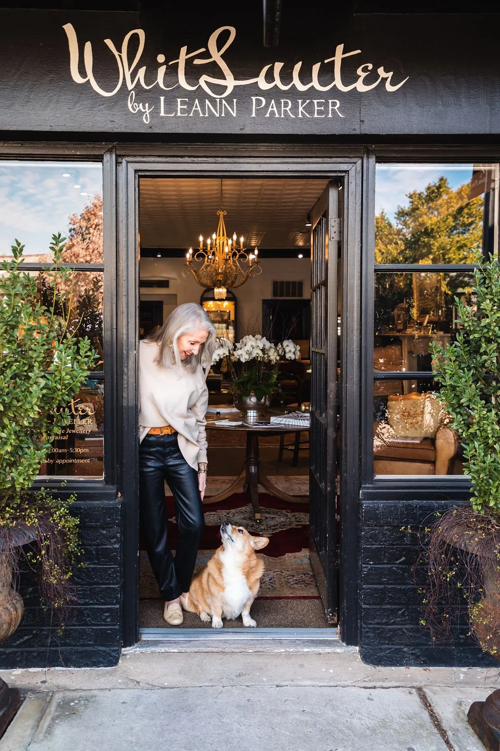 A woman with gray hair standing at the entrance of a boutique named 'White Sutter by Leann Parker,' looking down at her dog, a Pembroke Welsh Corgi, inside the shop. The boutique features a chandelier, a table with flowers, and a cozy interior visible through the glass doors.