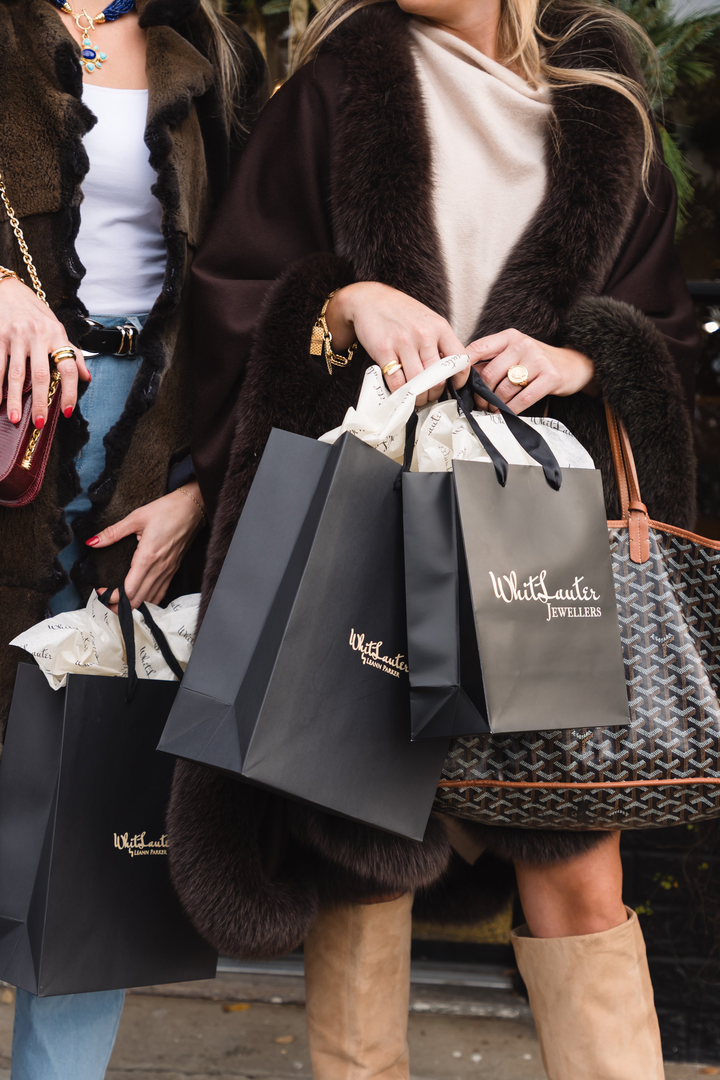 Women holding shopping bags from White Sauter Jewellers, dressed in winter coats with fur details, near outdoor Christmas decorations.