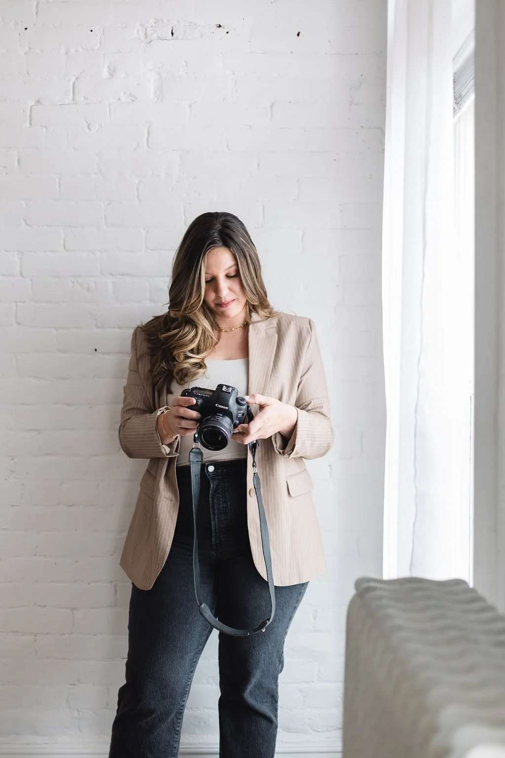 A woman with long hair wearing a beige blazer and dark jeans, holding a camera, standing in a bright room with white brick wall background and natural light from a window.