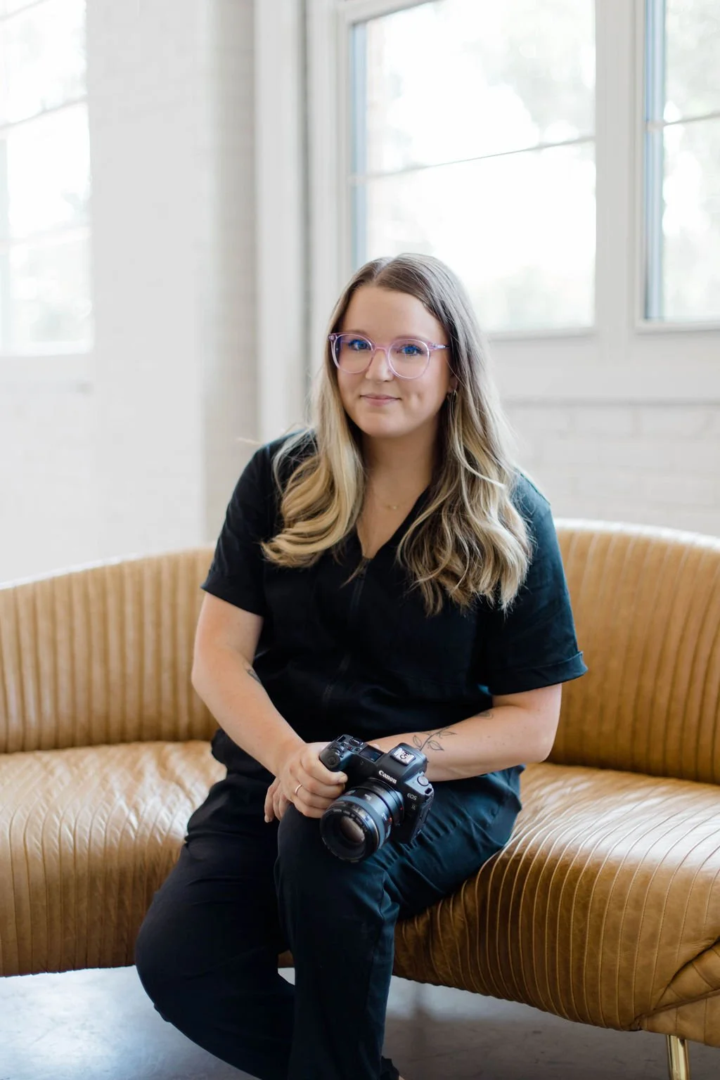A woman with glasses and long blonde hair sitting on a tan couch, holding a camera, in a room with large windows and white walls.