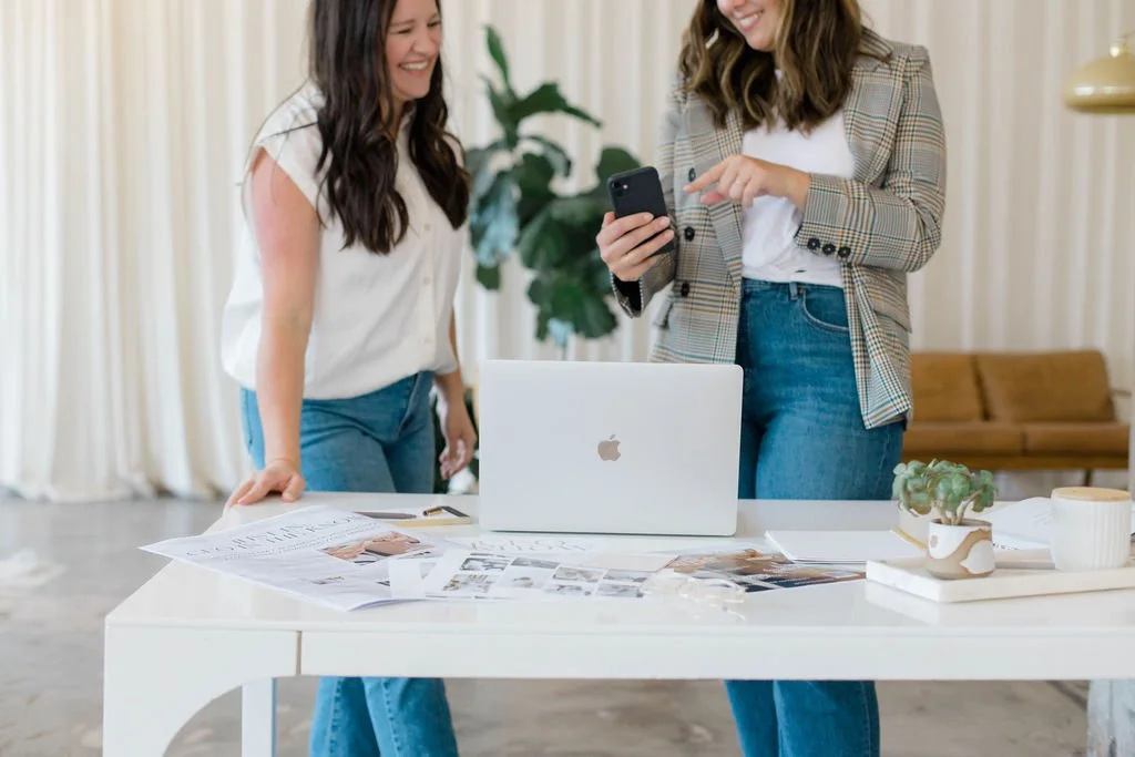 Two women standing at a desk, looking at a smartphone together, with a laptop and papers on the desk, in a bright room with a sofa and plants in the background.