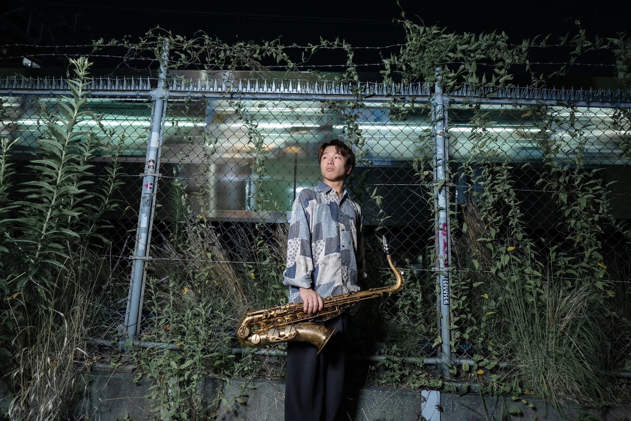 A young man with dark hair standing in front of a chain-link fence covered with climbing plants, holding a saxophone, at night.